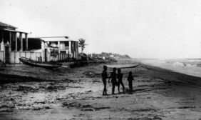 Schwarz weiß Foto. Vier Kinder, schemenhaft im Vordergrund, an einem Strand. Man sieht das Meer und einige Gebäude am Wasser.