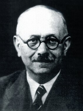 Portrait en noir et blanc de Marc Bloch, vêtu d'un costume, d'une chemise blanche, d'une cravate, portant des lunettes et une moustache